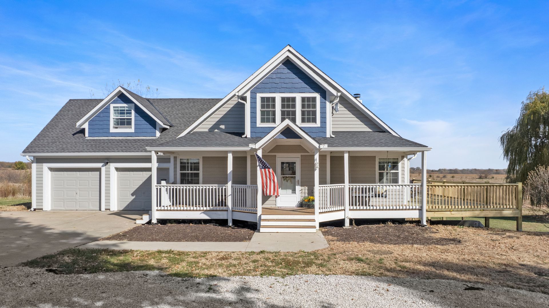 Front of home with blue paint, wrap around porch, and American Flag blowing in the wind. Gravel driveway leading up to paved driveaway.