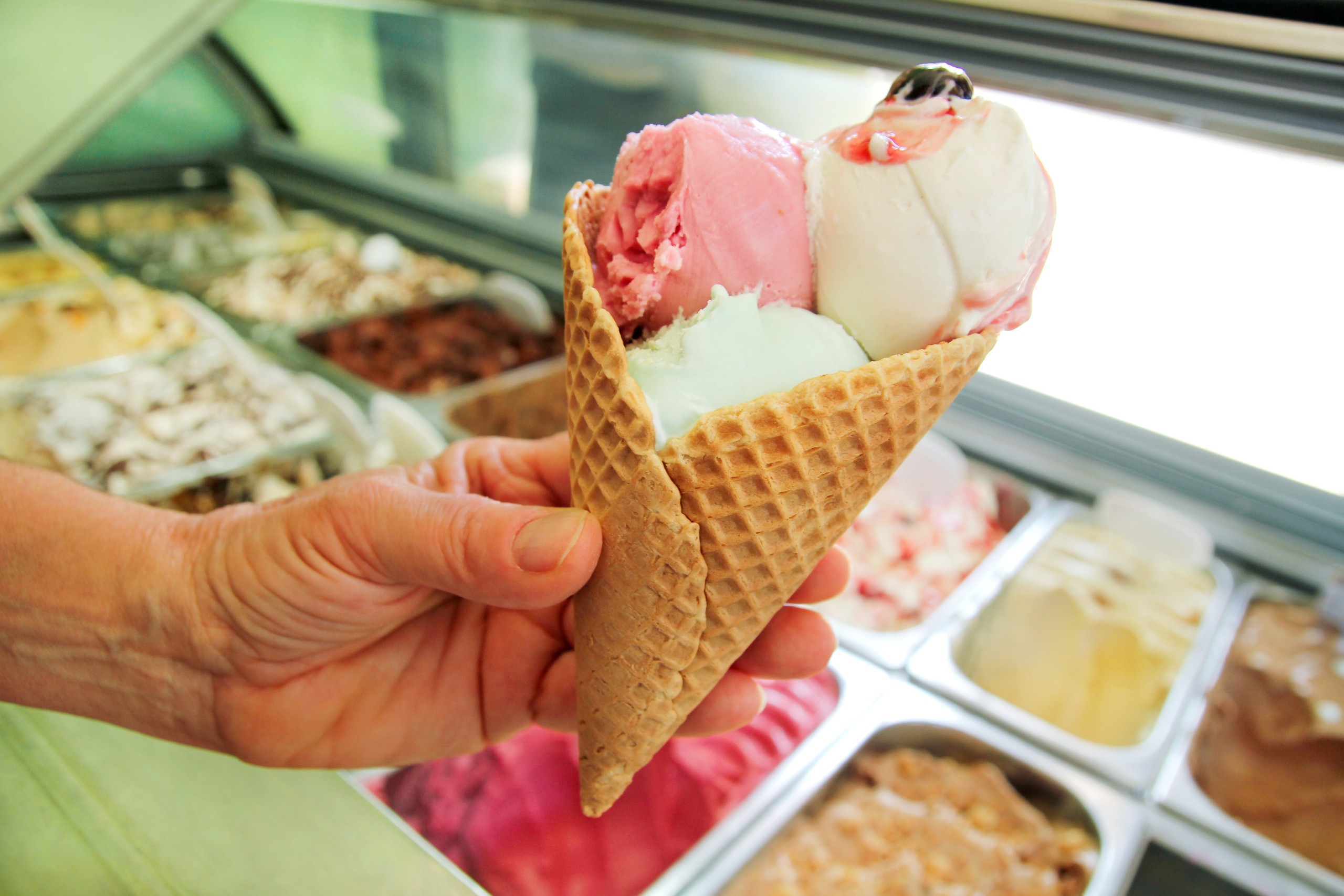 Female hand is holding a large strawberry, mint, vanilla with sour cherry ice cream in waffle cone. Ice cream fridge with steel service containers in background. 