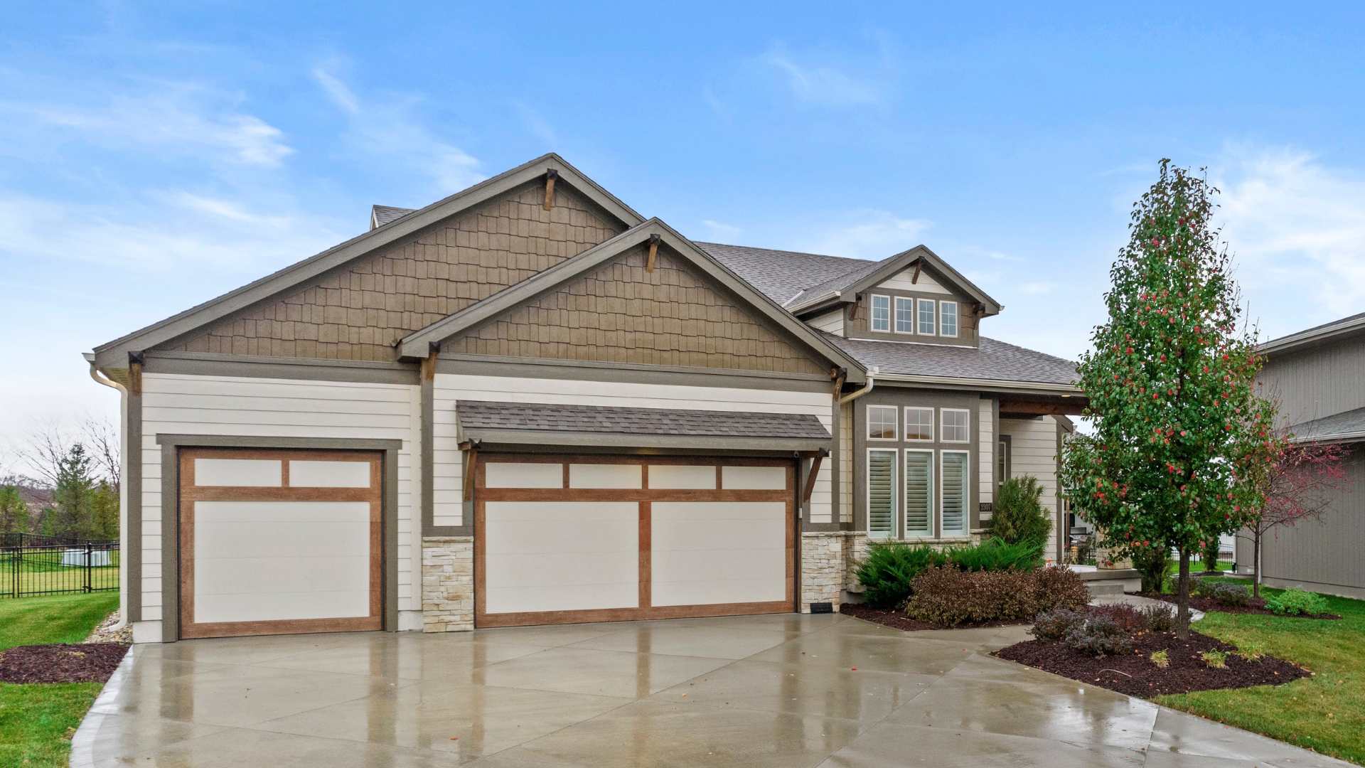 Front photo of a home with wet driveway from rain. Garage doors have light brown border and nice landscaping.